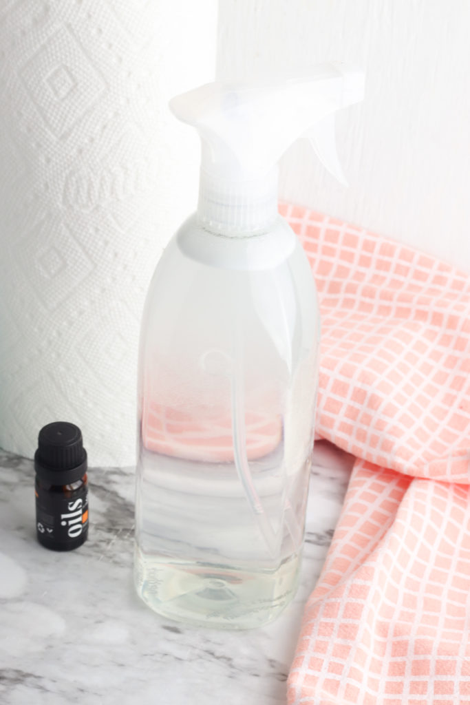 DIY window cleaner in a spray bottle next to paper towels, a red cloth and essential oil.