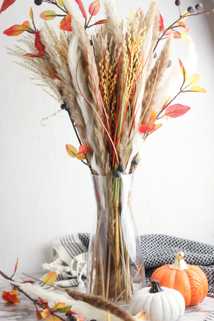 Pampas grass arrangement in a glass vase with fall decor and pumpkins.